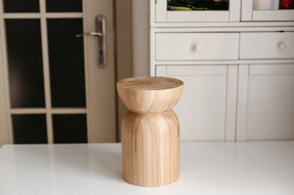 A small cylindrical wooden stool with a rounded top and chunky base, shown on a white table in a modern room with a white cabinet in the background.