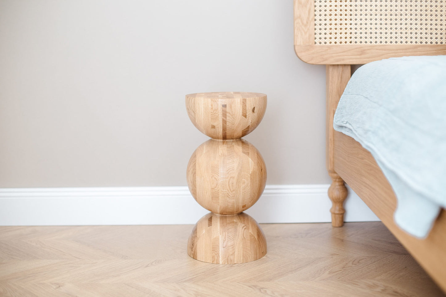 A handturned wooden bedside table with a natural oak finish, featuring a unique wave design, placed on a hardwood floor next to a bed with a blue bedsheet.